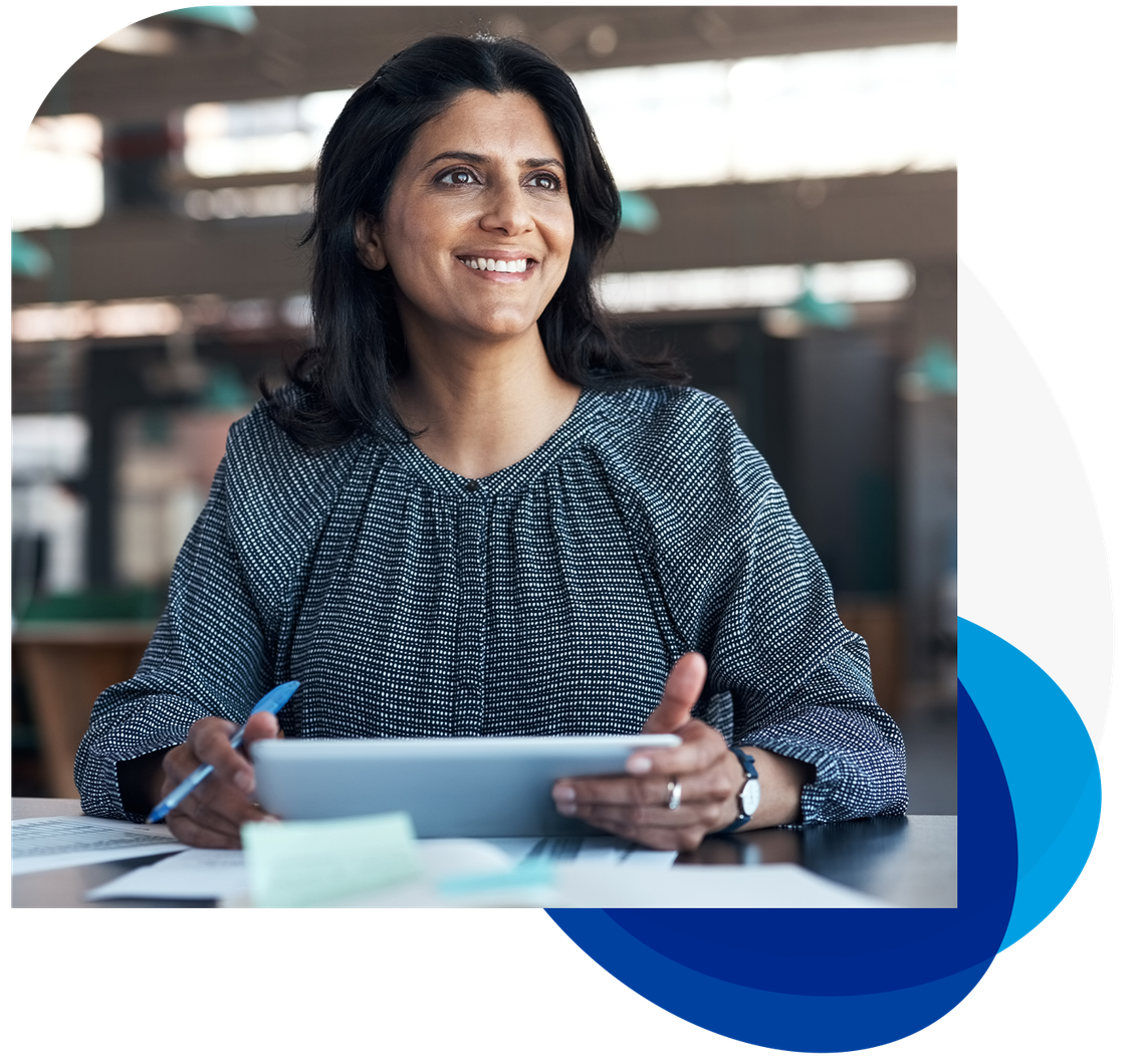 A woman is sitting at a desk, holding a tablet and smiling. She has a pen in her hand and papers scattered around her, with a modern office background visible.