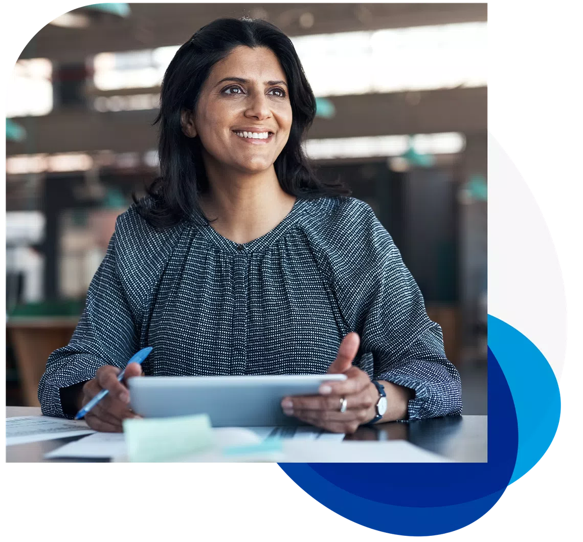 A woman is sitting at a desk, holding a tablet and smiling. She has a pen in her hand and papers scattered around her, with a modern office background visible.
