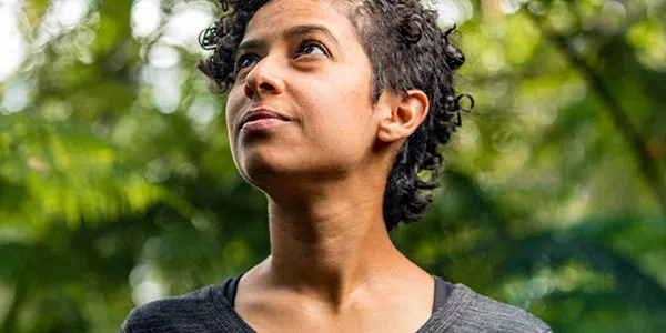 A person with short, curly hair gazes upward, appearing thoughtful or contemplative. They stand amidst lush greenery, with soft, natural lighting enhancing the serene atmosphere.