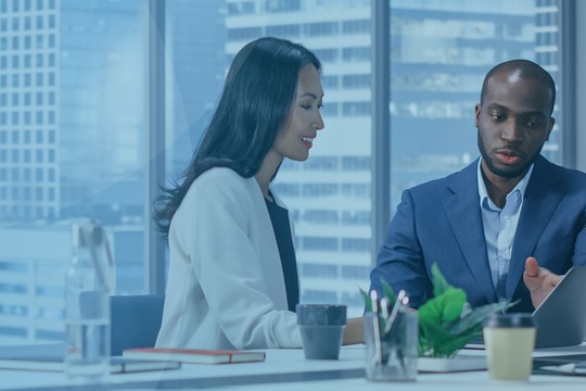 Two professionals engage in discussion at a modern, brightly lit office with large windows showcasing a city view. One person points at a laptop, while the other listens attentively.