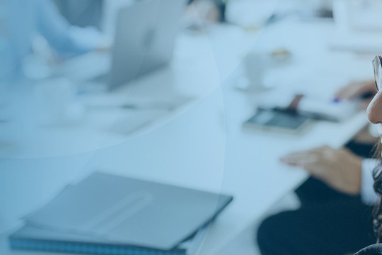 A woman with glasses smiles while seated at a conference table, engaging with others in a professional setting filled with laptops and notebooks. The background suggests a collaborative atmosphere.