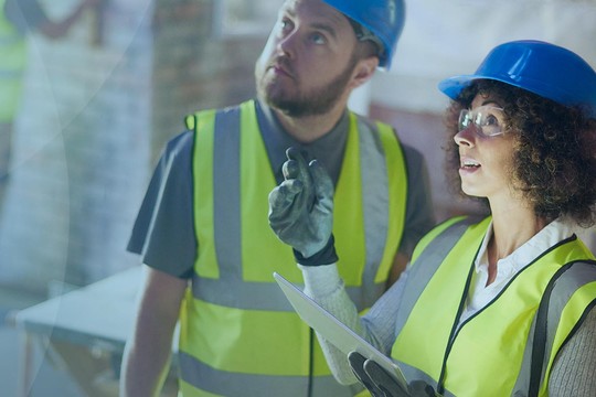 Two construction workers, wearing hard hats and high-visibility vests, are discussing plans while examining a building site, surrounded by unfinished walls and construction materials.
