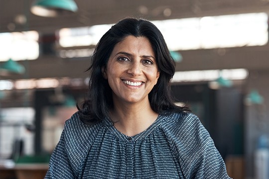 A woman with long black hair smiles warmly while sitting at a table. The modern, bright background features green pendant lights and a spacious interior, suggesting a welcoming atmosphere.
