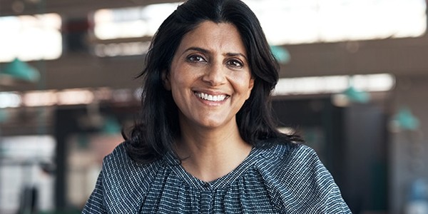 A woman with long black hair smiles warmly while sitting at a table. The modern, bright background features green pendant lights and a spacious interior, suggesting a welcoming atmosphere.