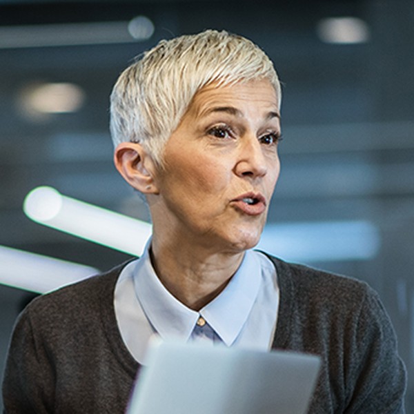 A woman with short hair holds a document while speaking animatedly to two men in a modern office setting. The room features large windows and contemporary lighting.