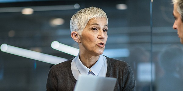 A woman with short hair holds a document while speaking animatedly to two men in a modern office setting. The room features large windows and contemporary lighting.