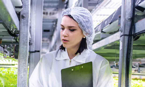 A woman in a white lab coat and hairnet examines data on a clipboard while standing between rows of plants in a greenhouse, surrounded by metal shelving.