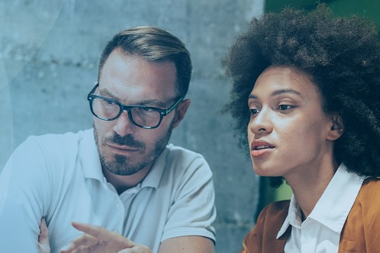 A man and a woman are engaged in a discussion, with the man appearing to analyze information, while the woman looks on attentively. The background features a modern, softly-lit space.