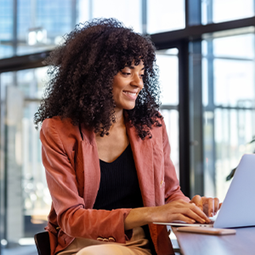 A woman with curly hair sits at a wooden table, smiling while working on a laptop. Large glass windows reveal a bright, modern office environment with greenery.