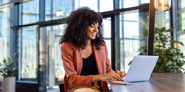 A woman with curly hair sits at a wooden table, smiling while working on a laptop. Large glass windows reveal a bright, modern office environment with greenery.