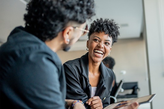 A woman with curly hair laughs while engaging with a man, who is partially visible. They appear to be in a bright, modern indoor space, suggesting a casual meeting or conversation.