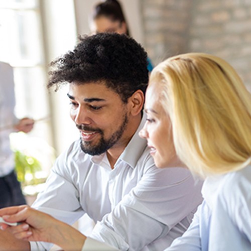 Two people are collaborating at a laptop, pointing at the screen and discussing. A third individual stands in the background, while a fourth is partially visible. The setting features a bright, modern workspace with stone walls.