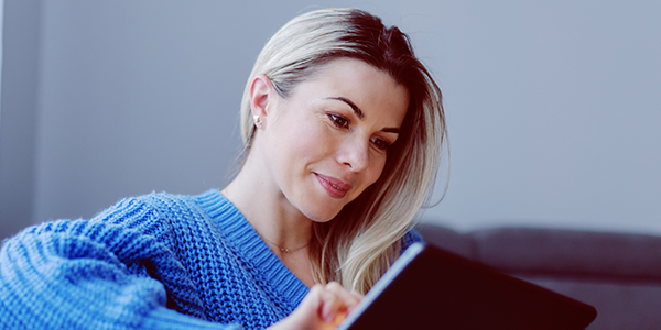 A woman wearing a blue sweater is attentively using a tablet while seated on a couch, with soft, natural light filtering through nearby curtains in a cozy living room setting.