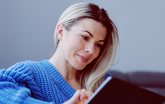 A woman wearing a blue sweater is attentively using a tablet while seated on a couch, with soft, natural light filtering through nearby curtains in a cozy living room setting.