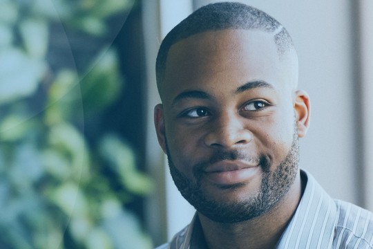 A man with short hair and a beard smiles while looking thoughtfully to the side. He's seated indoors with leafy green plants visible in the background, creating a calm atmosphere.
