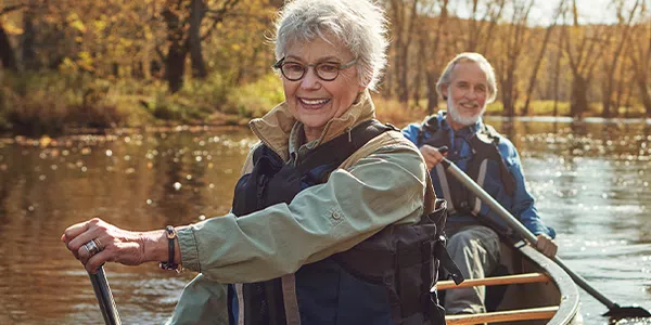 An elderly woman and man sit in a canoe on a calm river, paddling. Surrounding them are trees in autumn hues, creating a serene outdoor atmosphere accompanied by soft sunlight.