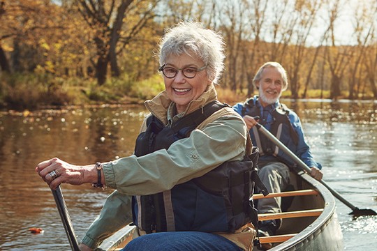 An elderly woman and man sit in a canoe on a calm river, paddling. Surrounding them are trees in autumn hues, creating a serene outdoor atmosphere accompanied by soft sunlight.