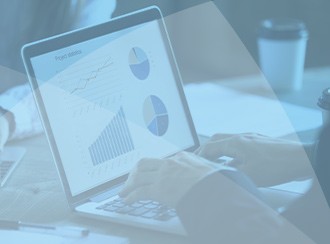 A laptop displays graphs and charts on a table. Hands are typing, analyzing data. A coffee cup is partially visible in the background, indicating a casual work environment.