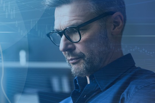 A man with glasses is focused on his work, surrounded by a blue-toned environment with abstract technological graphics, suggesting a digital or analytical context.