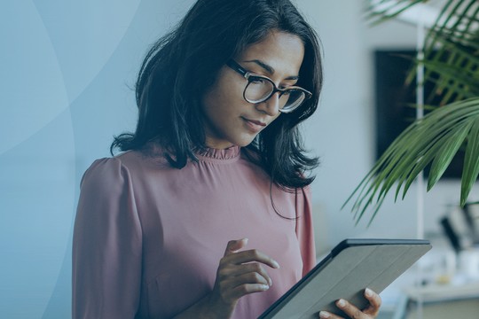 A woman with glasses holds a tablet, focused on its screen. She stands in a bright, modern office with a green plant nearby, conveying a productive atmosphere.