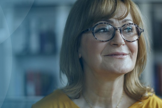 A woman with glasses smiles while looking off-camera. She sits in a cozy room, surrounded by shelves filled with books, suggesting a warm, inviting atmosphere.