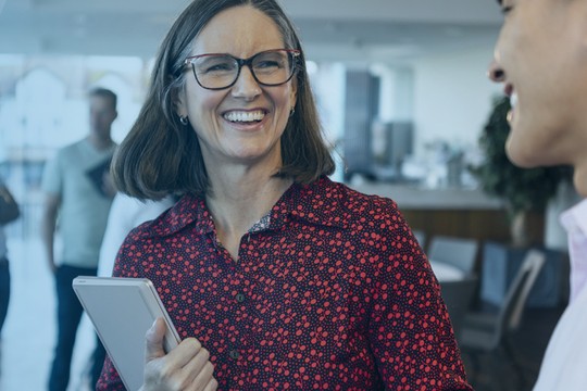 Two women are smiling and chatting, one wearing a hijab, while a man listens. They are in a modern, bright office environment, engaged in a friendly conversation.