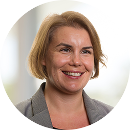A woman with short, blonde hair smiles confidently while wearing a gray blazer. The background is softly blurred, suggesting an indoor setting, possibly an office or professional environment.