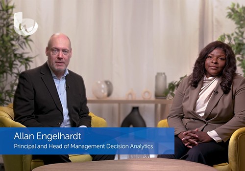 A man and a woman sit together on a yellow couch, engaged in conversation. The background features plants and decorative items, creating a professional but relaxed setting. 

Text: 
Allan Engelhardt  
Principal and Head of Management Decision Analytics