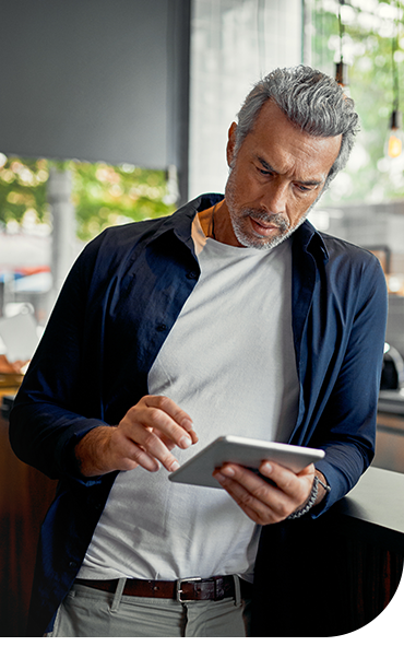 A man in casual attire stands by a counter, intently examining a tablet in his hands. Natural light filters through large windows, illuminating a modern café setting.