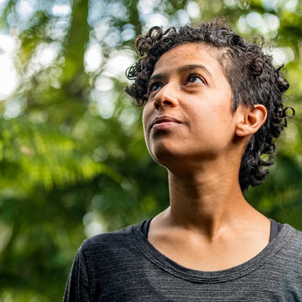 A woman with curly hair gazes upward, her expression thoughtful. She stands amidst vibrant green foliage, suggesting a serene, natural environment. Sunlight filters through the leaves, creating a tranquil atmosphere.