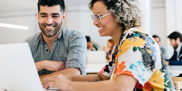 Two people are sitting at a table, happily engaging with a laptop. The woman types while the man observes with a smile. They are in a bright, modern workspace filled with other individuals.