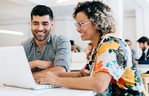Two people are sitting at a table, happily engaging with a laptop. The woman types while the man observes with a smile. They are in a bright, modern workspace filled with other individuals.