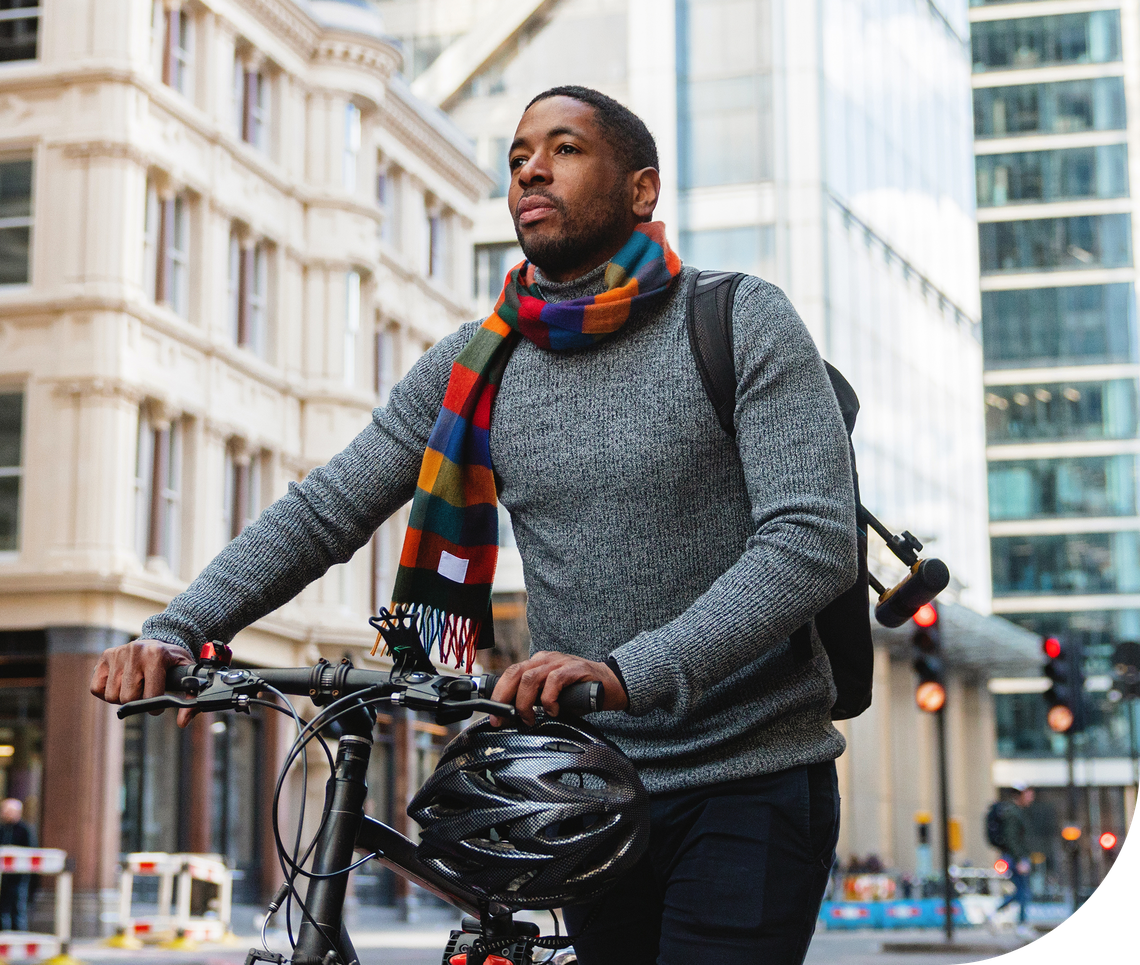 A man wearing a colorful scarf walks alongside his bicycle in an urban setting, surrounded by modern buildings and traffic signals, suggesting a lively city atmosphere.