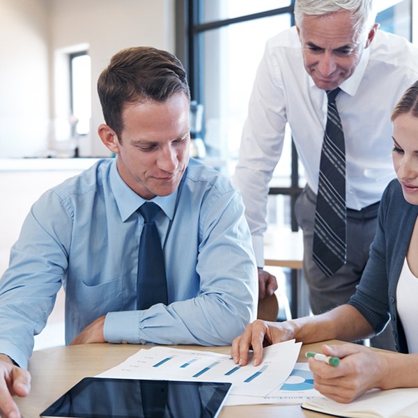 Three professionals are collaborating at a table. One man and one woman are analyzing a document with graphs, while another man observes. The setting appears to be a modern office.