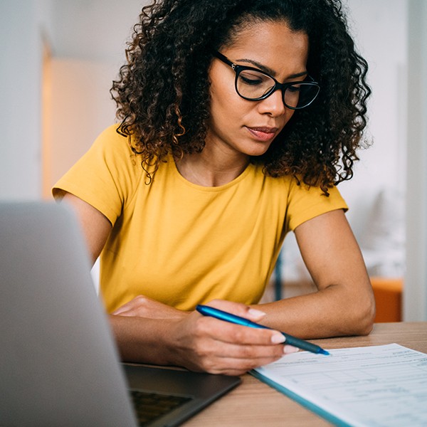 A woman with curly hair, wearing a yellow shirt, writes on a form with a blue pen while sitting at a desk with a laptop in a bright, minimalist room.