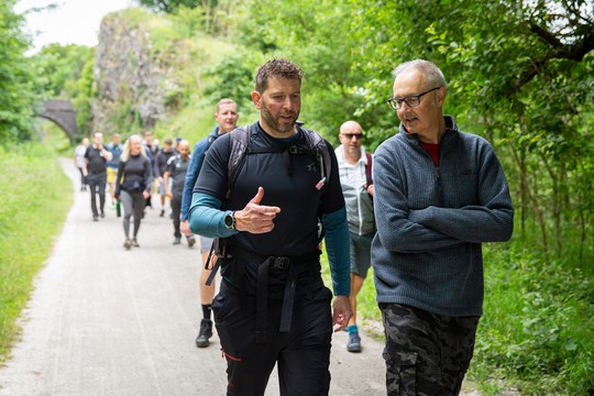 Two men walk on a gravel path, engaged in conversation. Surrounding them is a group of hikers, with greenery and a stone bridge in the background.