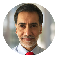 A man with a neatly trimmed beard smiles directly at the viewer. He wears a light blue shirt and a red tie. The background suggests a blurred indoor setting.