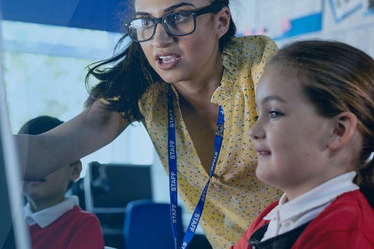 A teacher interacts with a student at a computer in a classroom. The teacher points at the screen, while another student listens attentively. The environment features colorful educational materials and desks.
