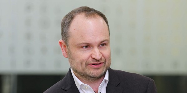 A man with short hair and a slight beard is speaking, conveying information. He is dressed in a dark suit with a white shirt, and a blurred modern background suggests a professional setting.
