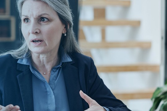 A woman with long hair gestures expressively while engaged in a conversation. She wears a dark blazer over a light blue shirt, seated in a bright room with wooden stairs in the background.