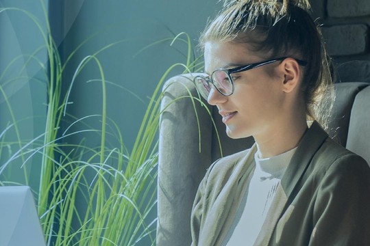 A woman with glasses sits on a couch, intently working on a laptop. Sunlight illuminates a green plant beside her in a modern, warmly lit indoor setting.