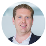 A man with light hair and a friendly smile is wearing a white shirt and a dark blazer. He is positioned against a softly blurred indoor background, likely an office setting.