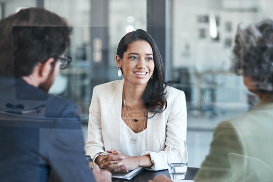 A woman in a white blazer smiles while seated at a conference table. She engages with two colleagues, one wearing glasses, in a bright, modern office setting. A glass of water is nearby.