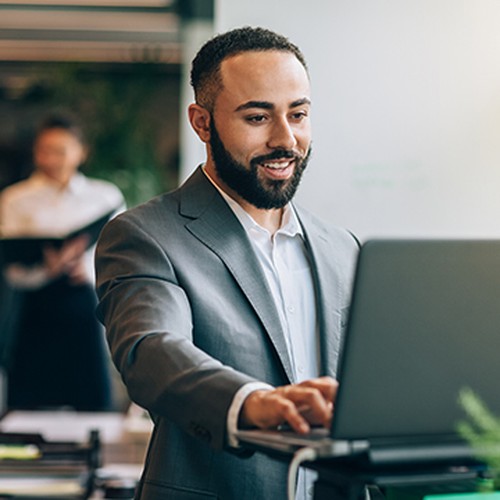 A man in a suit smiles while engaging with a laptop on a stand. In the background, a person is seen holding documents in a modern office setting filled with plants and desks.