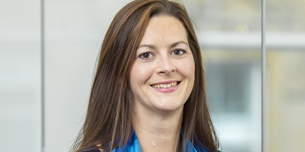 A woman with long, brown hair smiles while wearing a blue scarf and a dark top. She is positioned in front of a blurred, bright background.