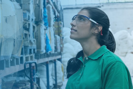 A woman wearing safety glasses and a green shirt examines stacks of compressed materials in a warehouse, surrounded by colorful packaging and other storage items, suggesting a recycling or manufacturing environment.