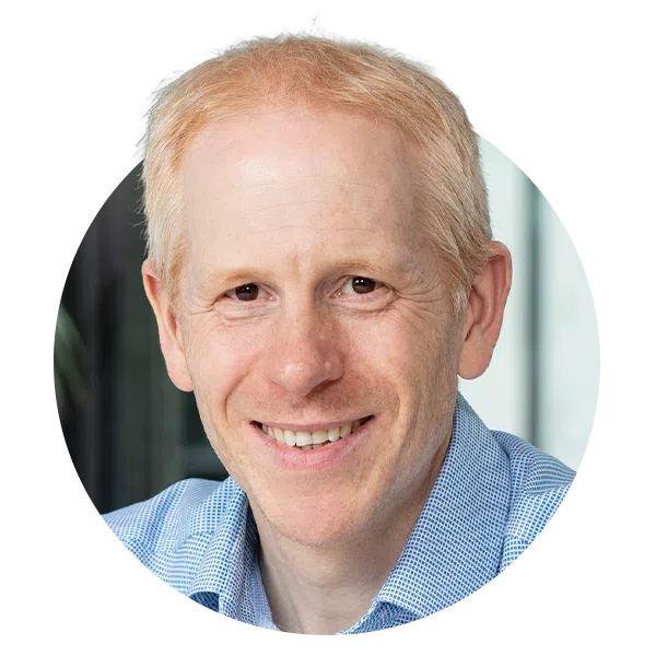 A smiling man with light blonde hair wears a blue checkered shirt. He is positioned in front of a blurred, modern interior, suggesting a professional environment.