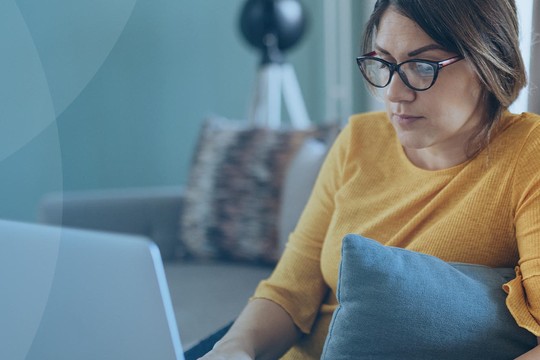 A woman in a yellow sweater sits on a sofa, focused on her laptop. She holds a blue cushion and is surrounded by a modern, softly lit living room with decorative elements.