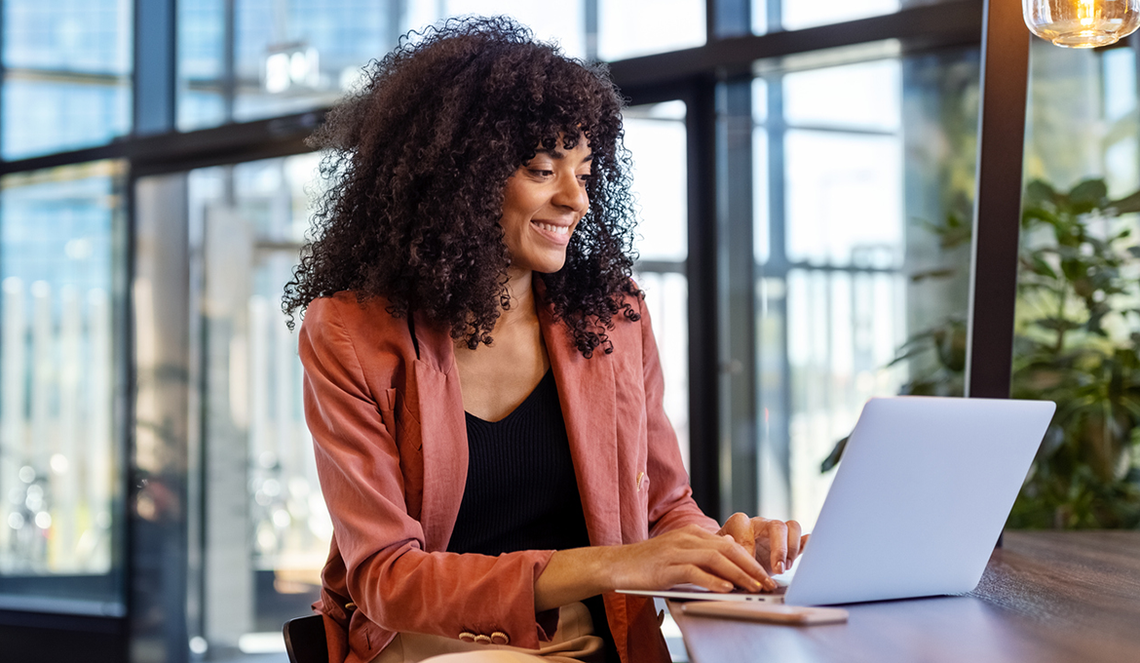 A woman with curly hair is smiling while typing on a laptop in a modern indoor setting, characterized by large windows and plants, suggesting a bright and inviting workspace.