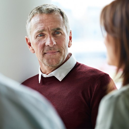 A middle-aged man wearing a maroon sweater listens attentively, engaging in conversation with two others in a well-lit indoor setting, with soft, blurred backgrounds.